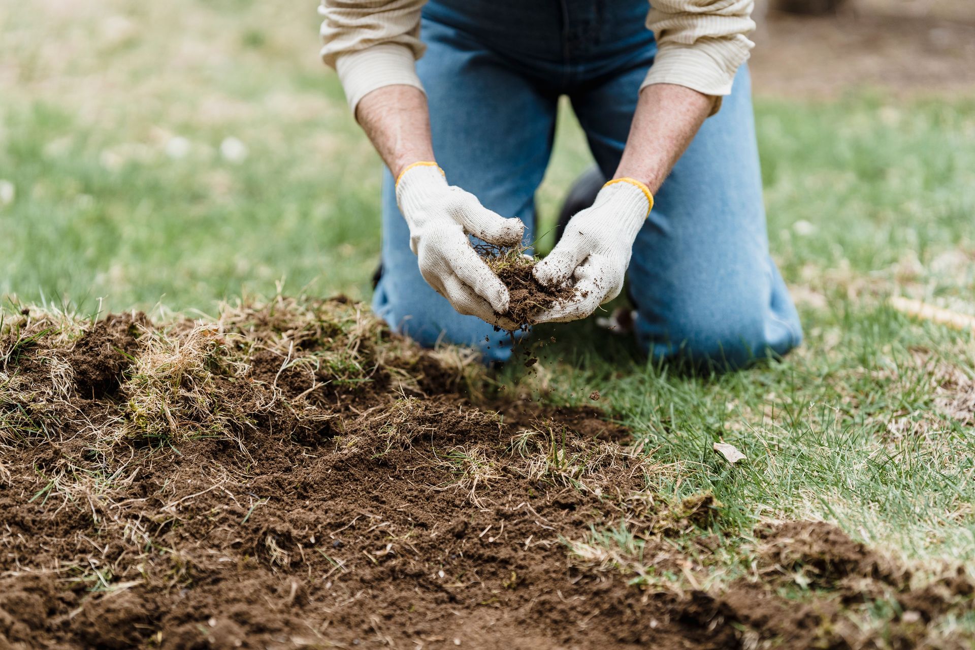 Crop unrecognizable gardener in casual clothes and gloves sitting on grassy ground while preparing soil for planting in garden